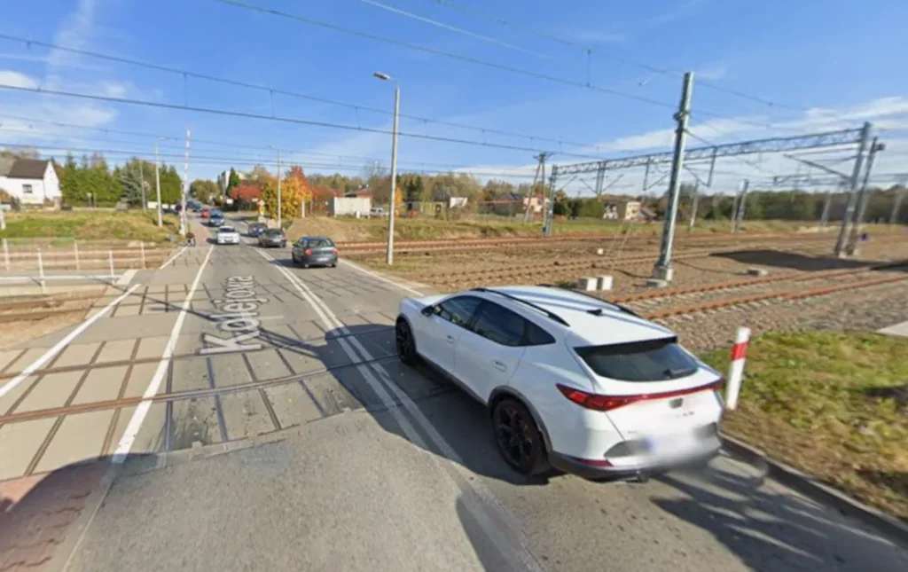 Obraz przedstawia widok z poziomu ulicy na wielotorowy przejazd kolejowy (level crossing) pod bezchmurnym niebem, patrząc wzdłuż torów. Po prawej stronie na poboczu zaparkowany jest biały crossover SUV, częściowo zasłani